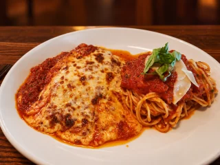 Chicken parmigiana with browned melted cheese and tomato sauce beside spaghetti on a white plate.