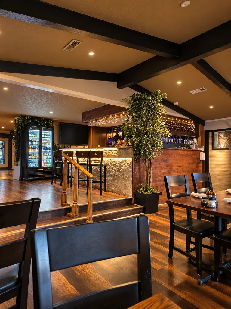 Dark wood tables and chairs facing the raised bar, with exposed ceiling beams, warm lighting and a tall potted tree beside the steps.