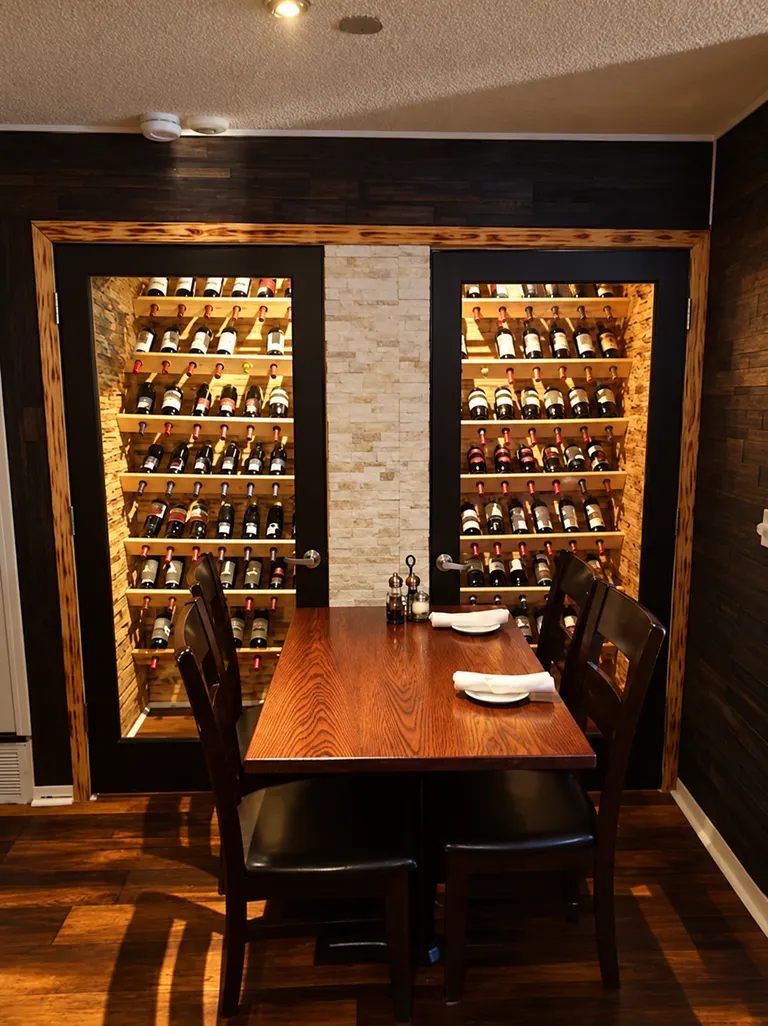 A wooden dining table and dark chairs in front of two illuminated wine racks behind glass doors, divided by a pale stone column.